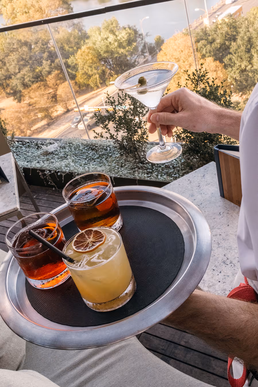 A person holds a martini while carrying a tray with three cocktails on an outdoor patio overlooking trees and a road.