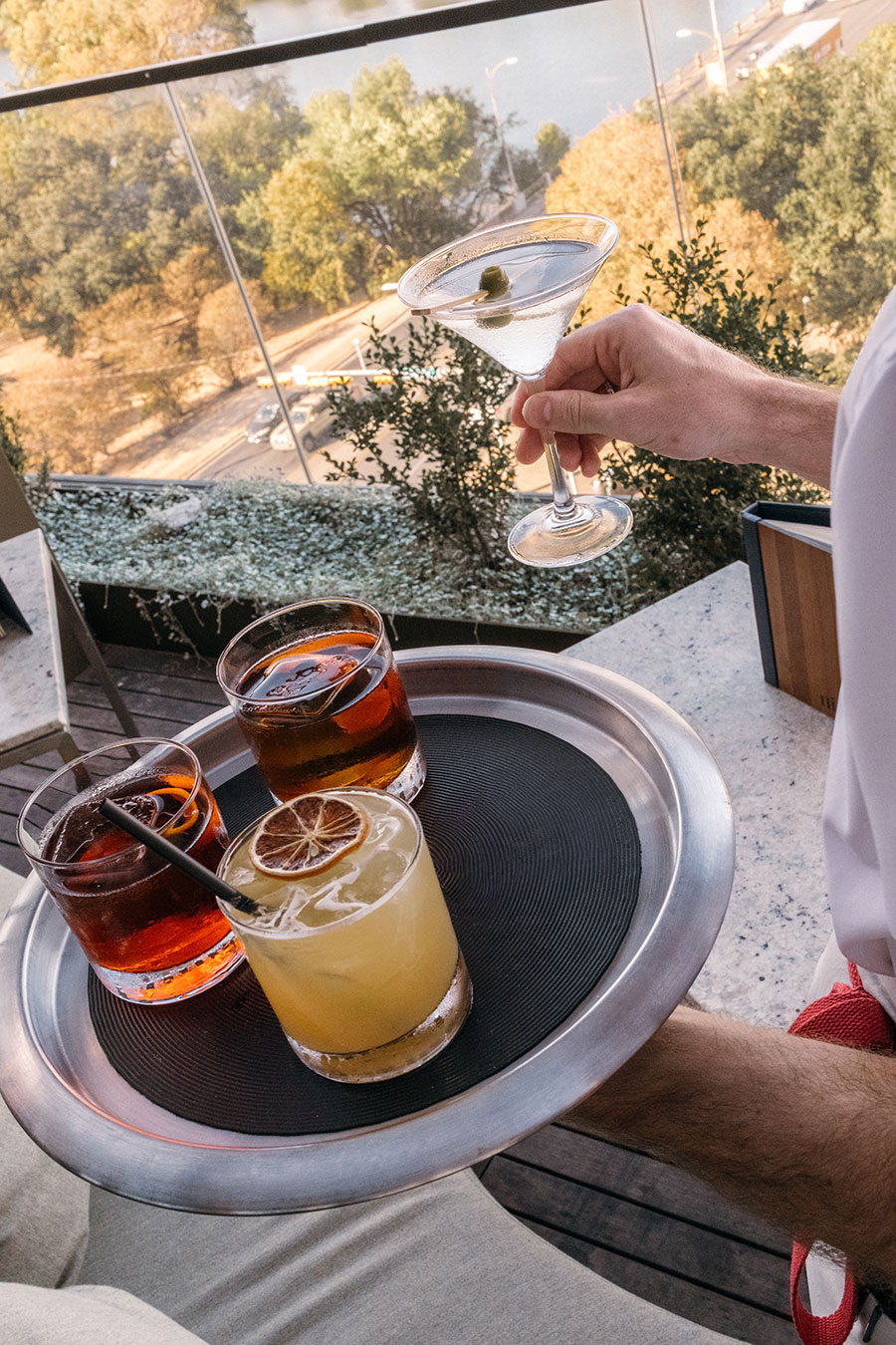 A person holds a martini while carrying a tray with three cocktails on an outdoor patio overlooking trees and a road.