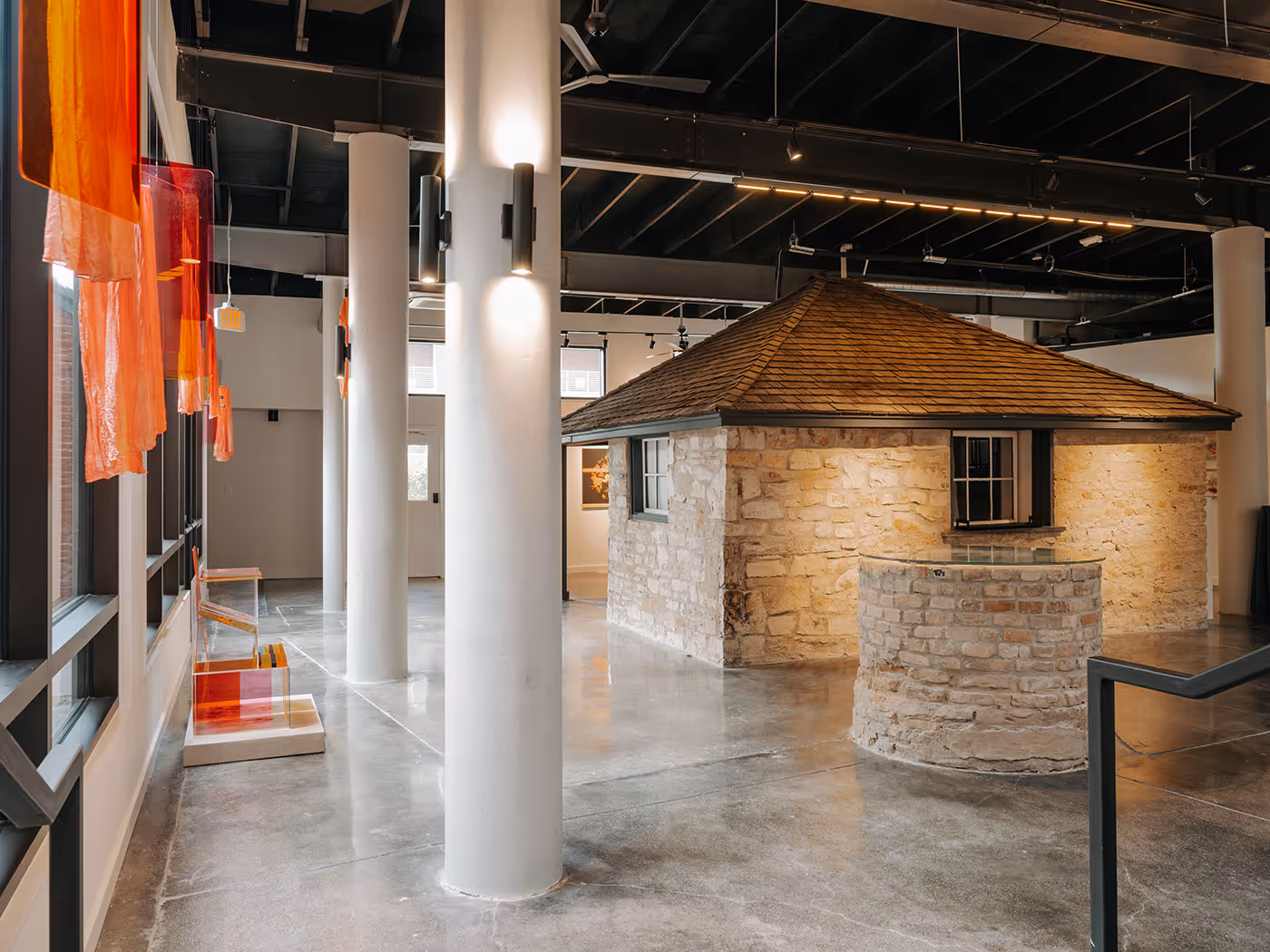 A small stone house with a wooden roof is displayed inside a modern gallery with large windows and art installations.