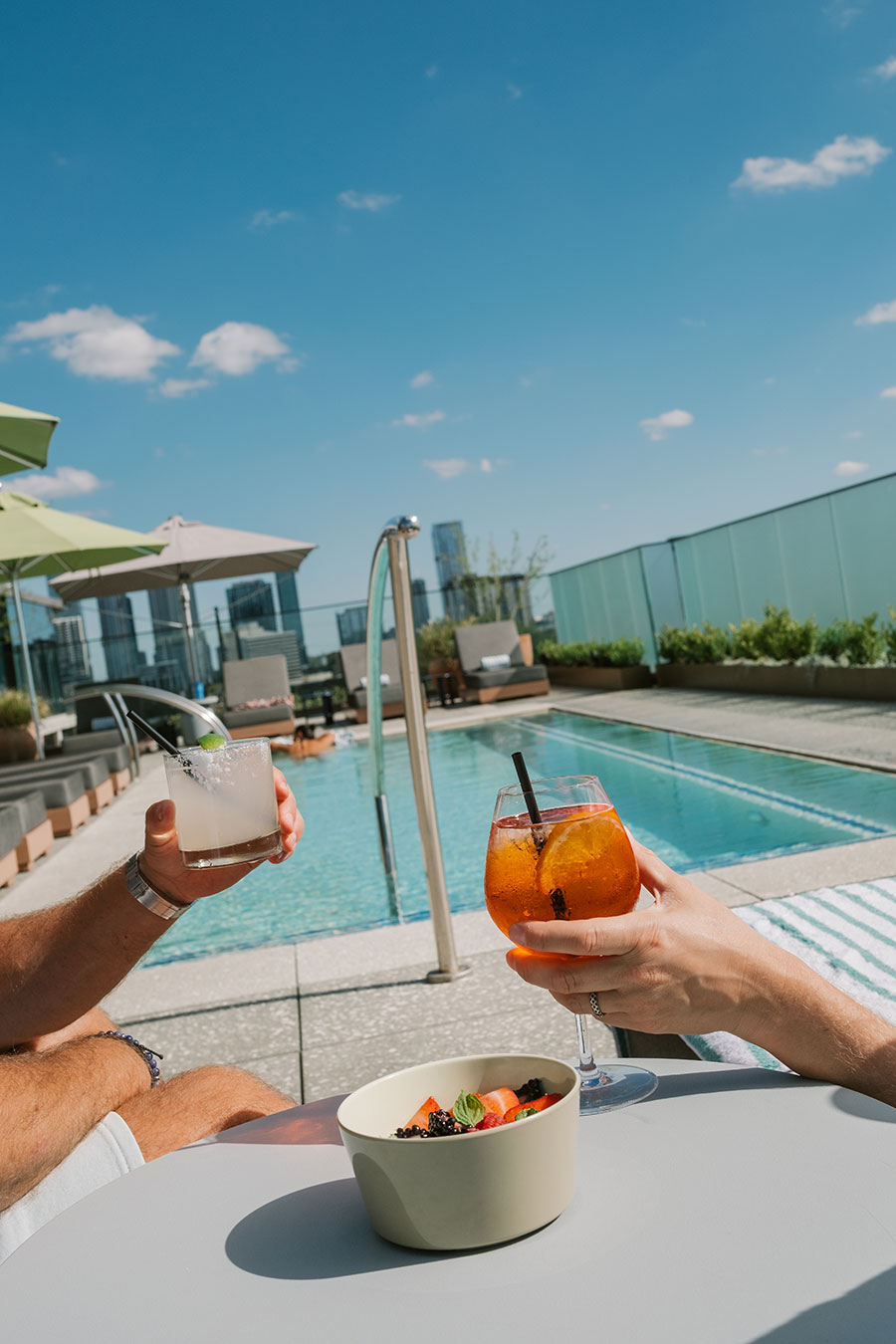 Two people hold cocktails by a rooftop pool with a bowl of fruit on a table under a clear blue sky.