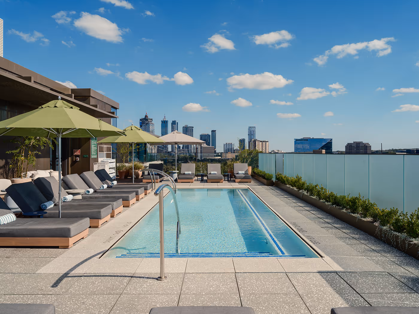 Rooftop pool with lounge chairs, umbrellas, and a glass railing overlooking a city skyline on a clear day.