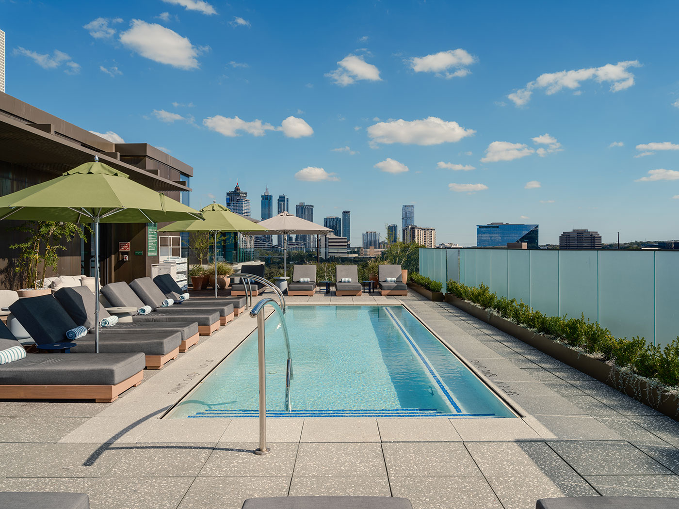 Rooftop pool with lounge chairs, umbrellas, and a glass railing overlooking a city skyline on a clear day.