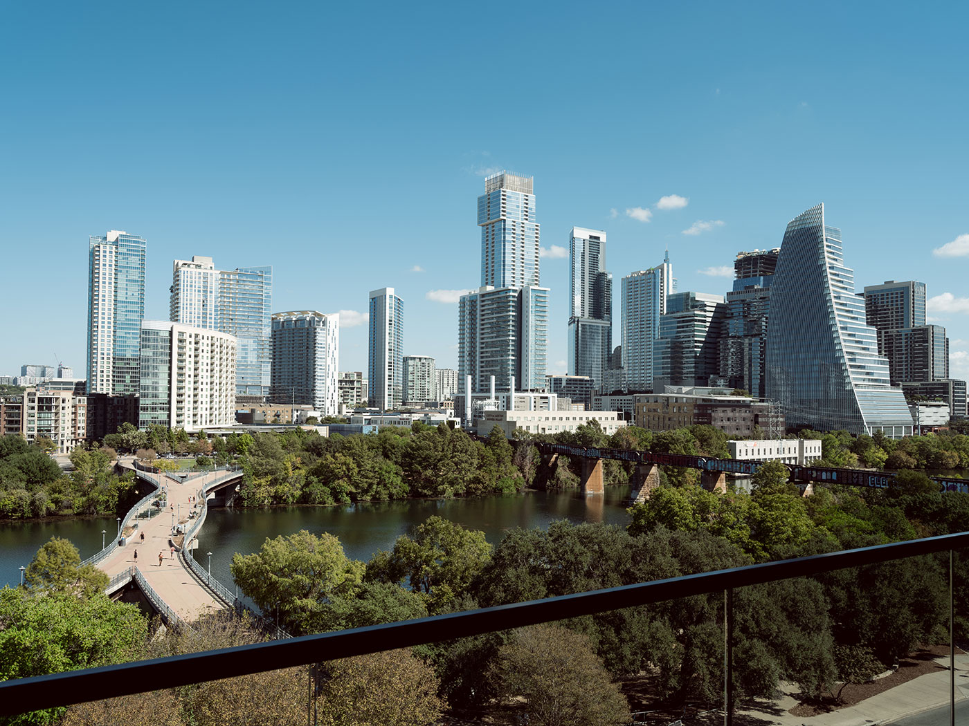 Downtown Austin skyline with modern high-rise buildings, a pedestrian bridge, and trees bordering a river under a clear sky.