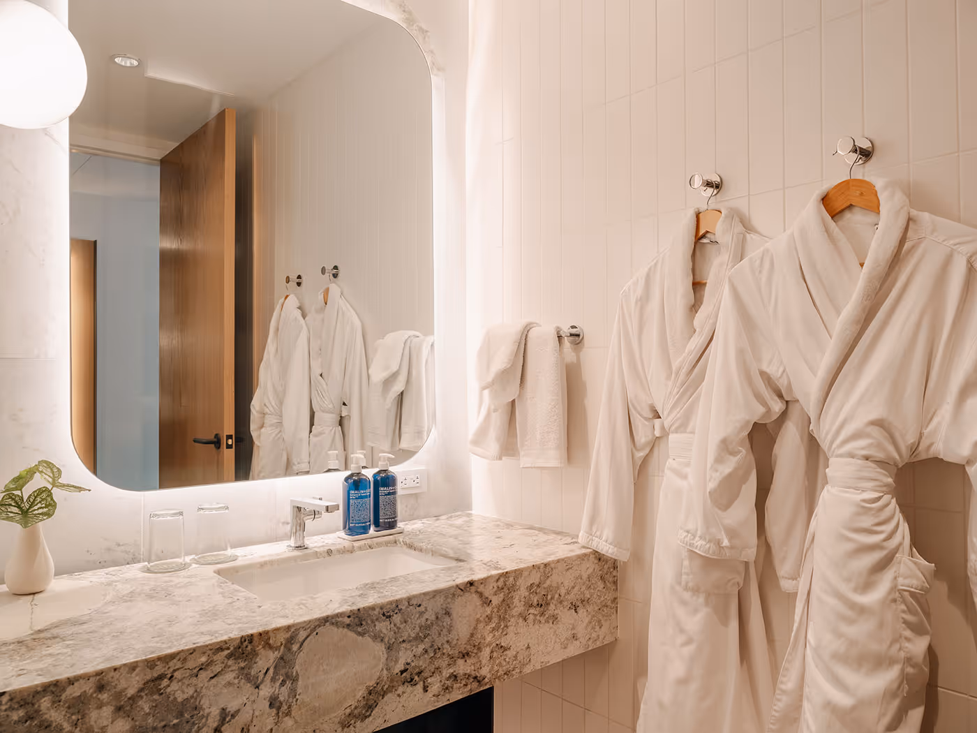 Modern bathroom with two white robes on hooks, a marble countertop, towels, toiletries, and a large backlit mirror.