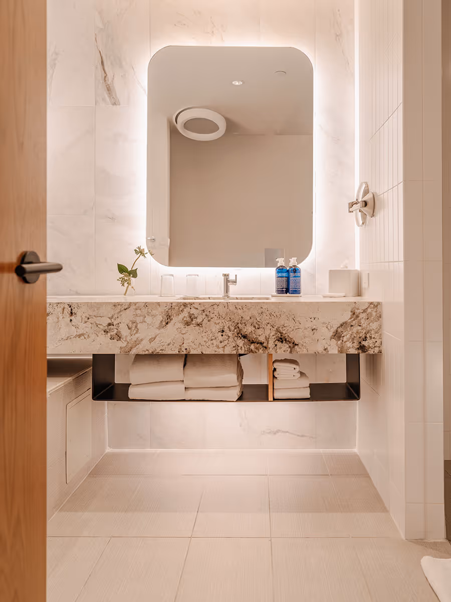 Modern bathroom with a backlit mirror, marble countertop, towels, toiletries, and minimalistic decor.