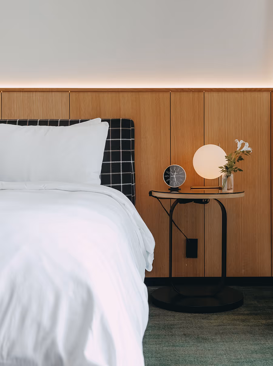 A neatly made bed beside a small table with a clock, round lamp, and vase of flowers against a wood-paneled wall.