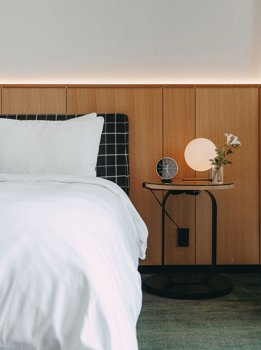 A neatly made bed beside a small table with a clock, round lamp, and vase of flowers against a wood-paneled wall.