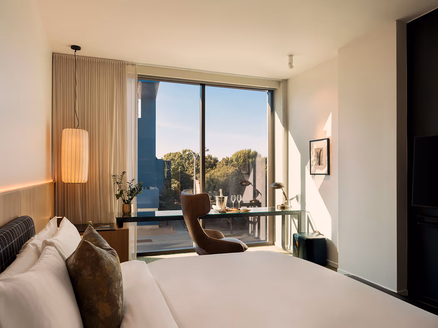 Modern hotel room with a bed, desk, chair, and large window overlooking trees and an outdoor balcony.