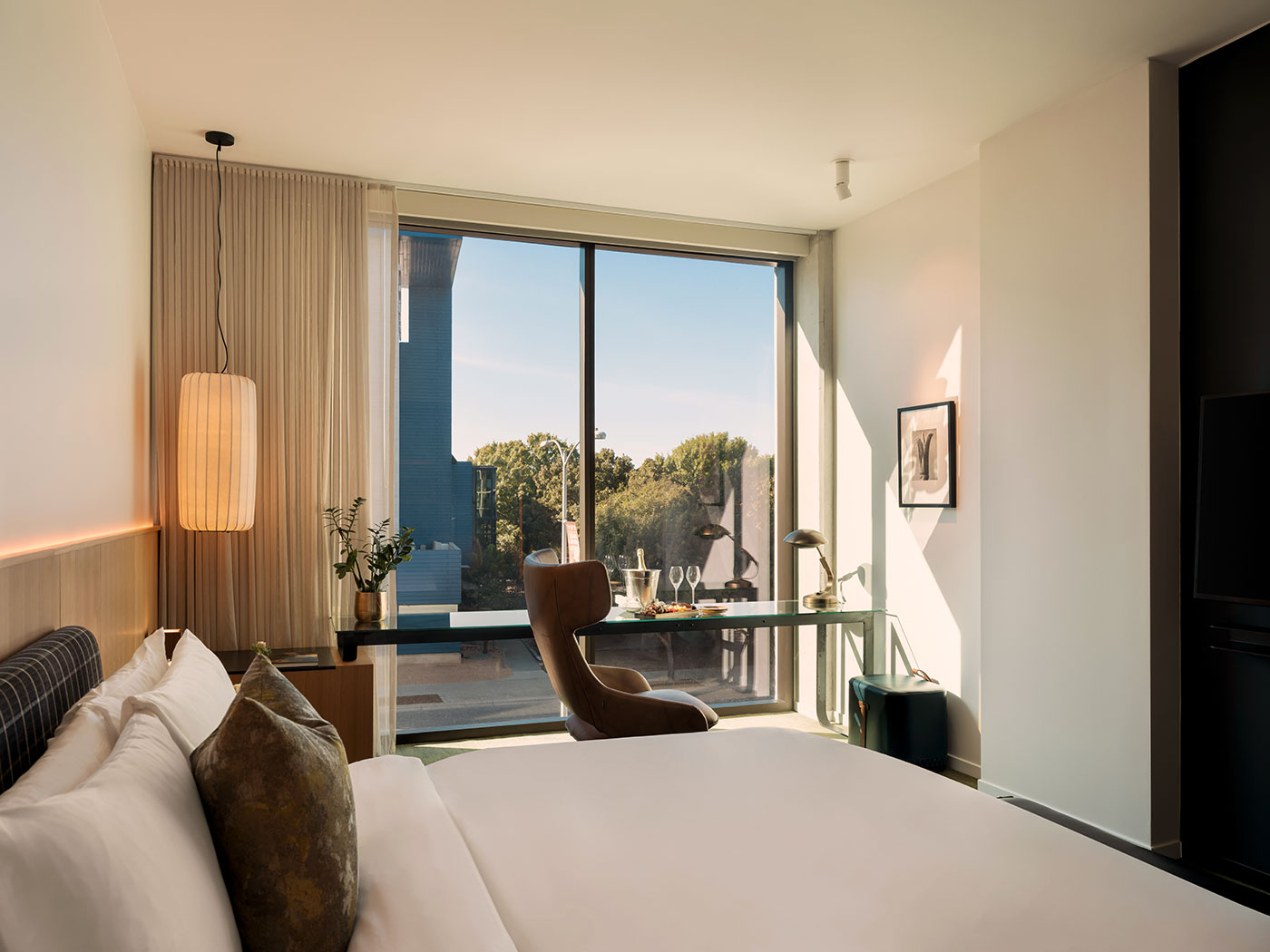 Modern hotel room with a bed, desk, chair, and large window overlooking trees and an outdoor balcony.