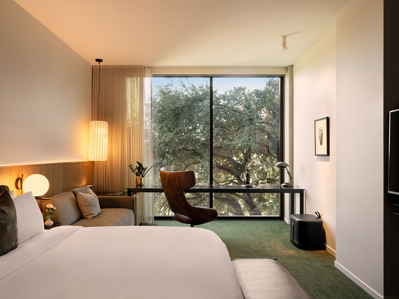 Modern hotel room with a bed, desk, chair, and large window overlooking leafy trees outside.