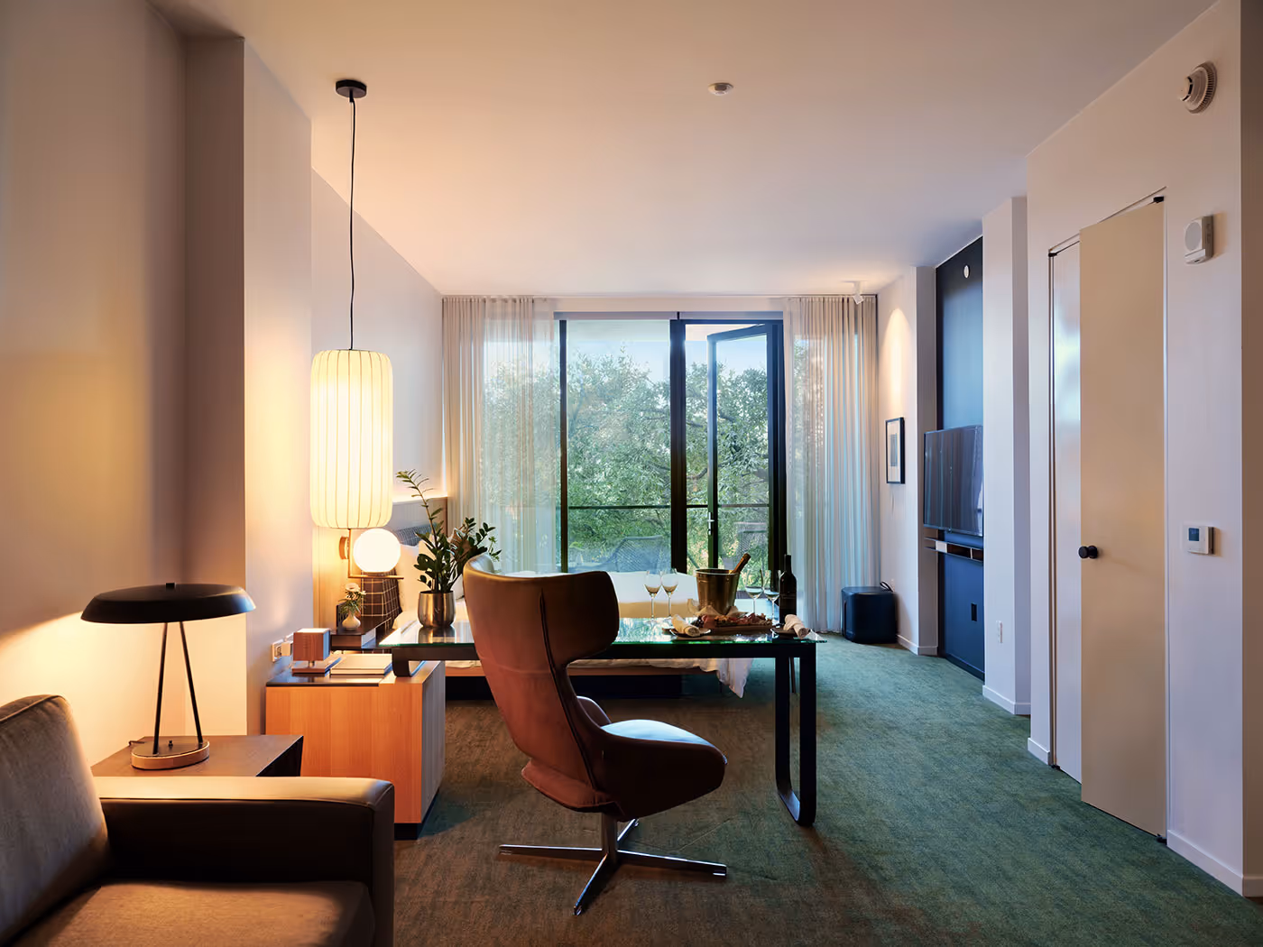 Modern living room with a desk, chair, open balcony door, and large window overlooking green trees.