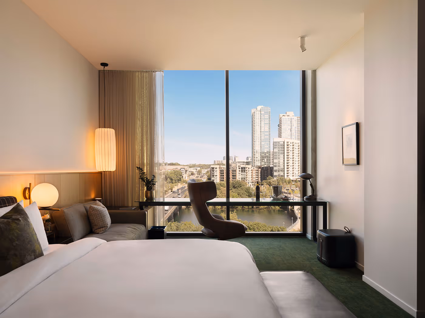 Modern hotel room with large window, cityscape view, bed, desk, chair, and contemporary decor.