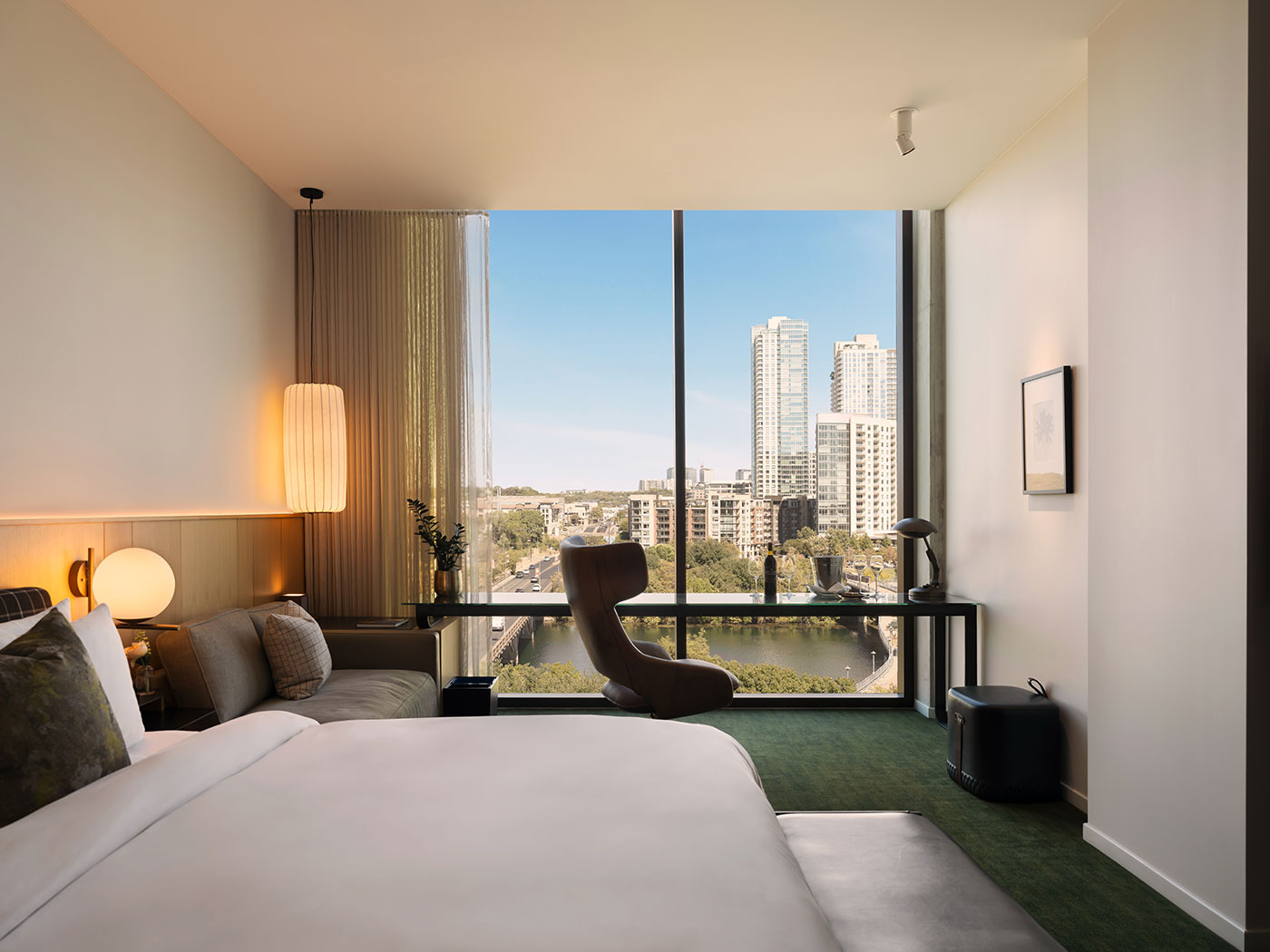 Modern hotel room with large window, cityscape view, bed, desk, chair, and contemporary decor.