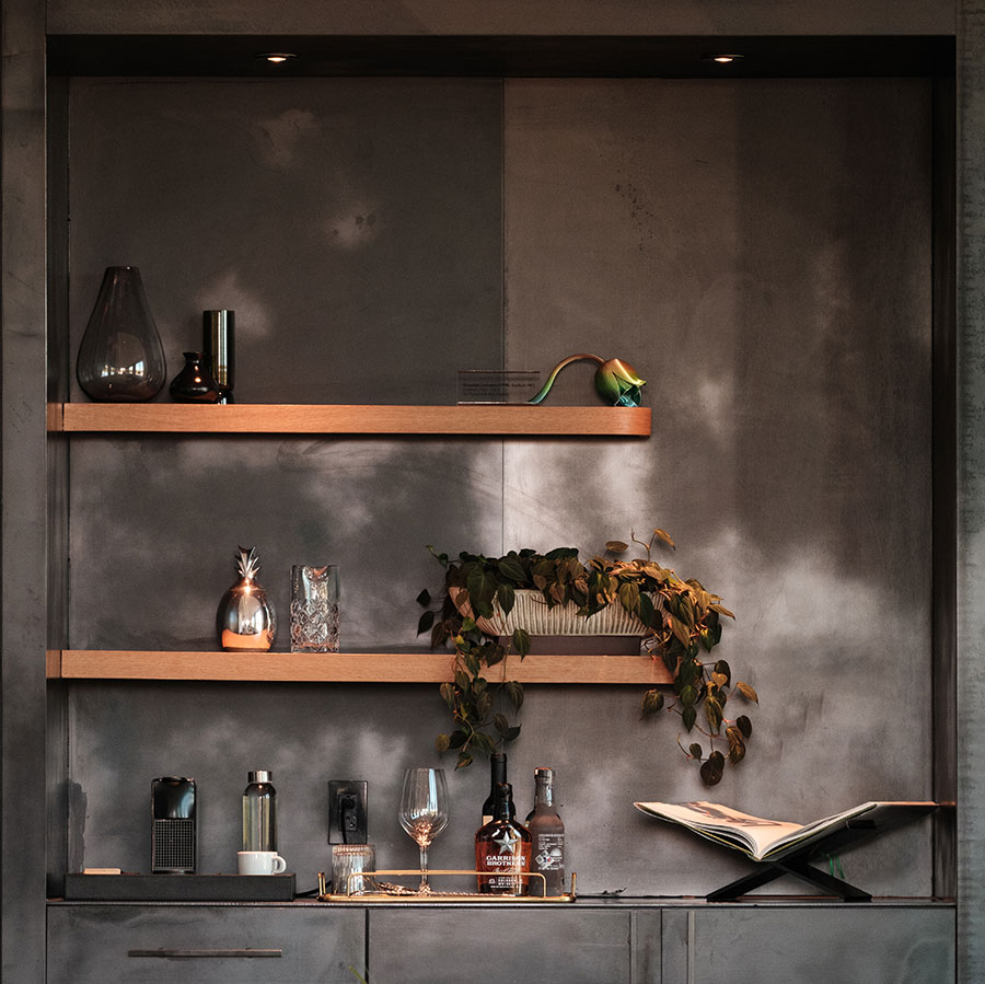 Modern shelves with decorative objects, bottles, a plant, glassware, and an open book against a gray wall.