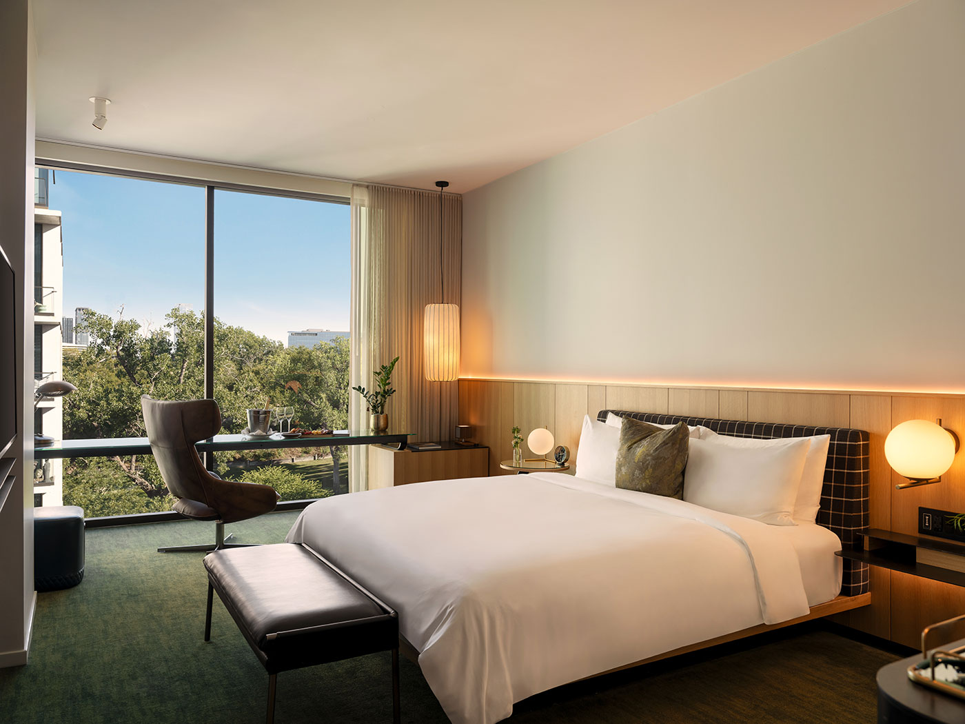 Modern hotel room with a large bed, desk, chair, and floor-to-ceiling window overlooking trees and buildings.