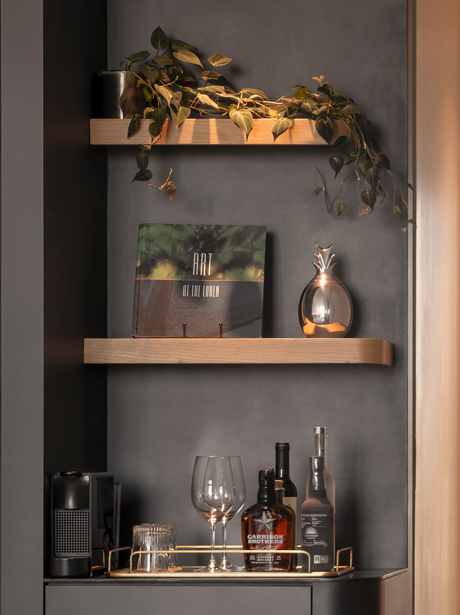 Two wooden shelves hold a potted plant, a book, and decor; below is a tray with glassware and bottles on a dark wall.
