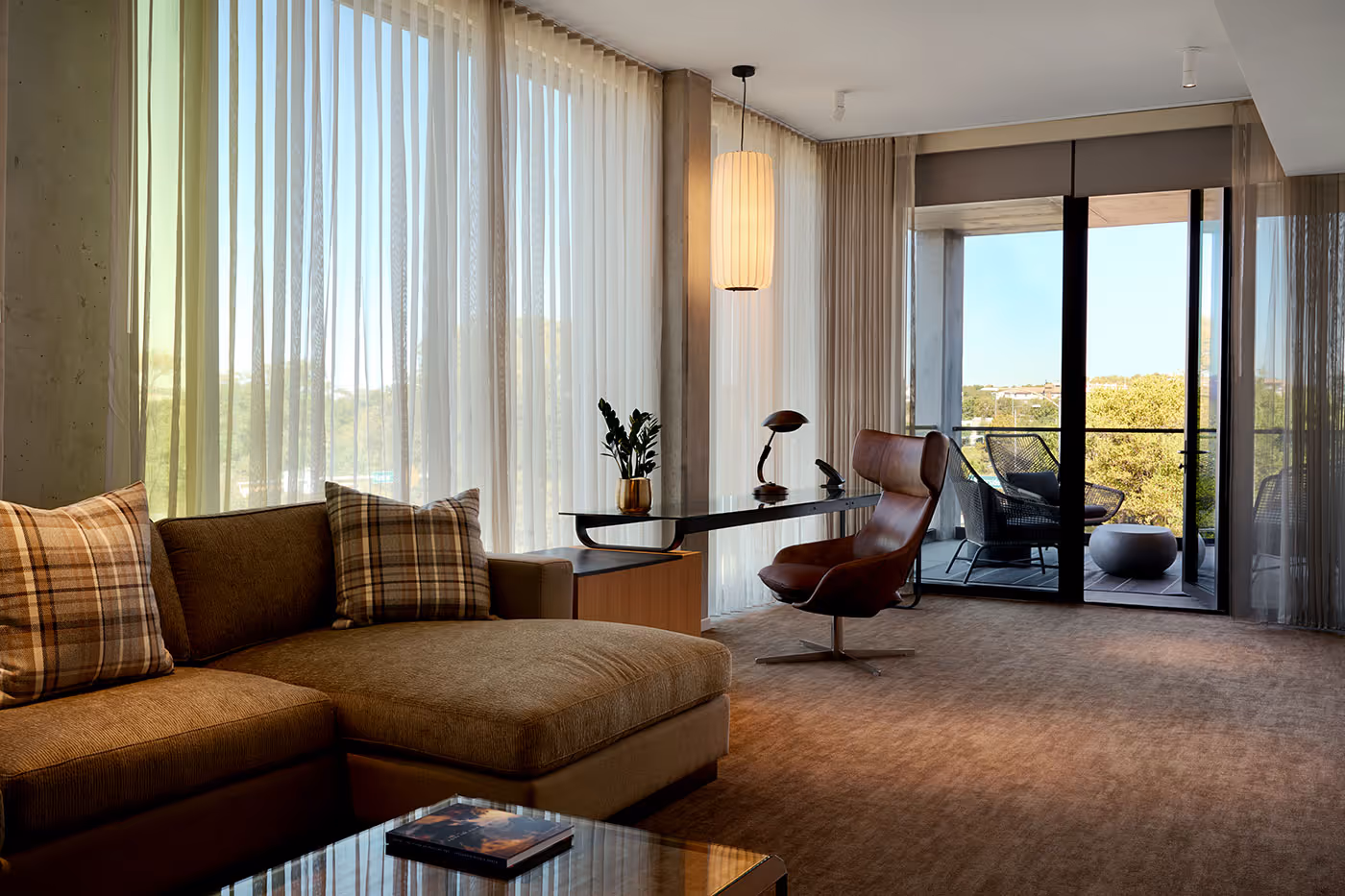 Modern living room with a sectional sofa, desk, lounge chair, large windows, and a balcony view.