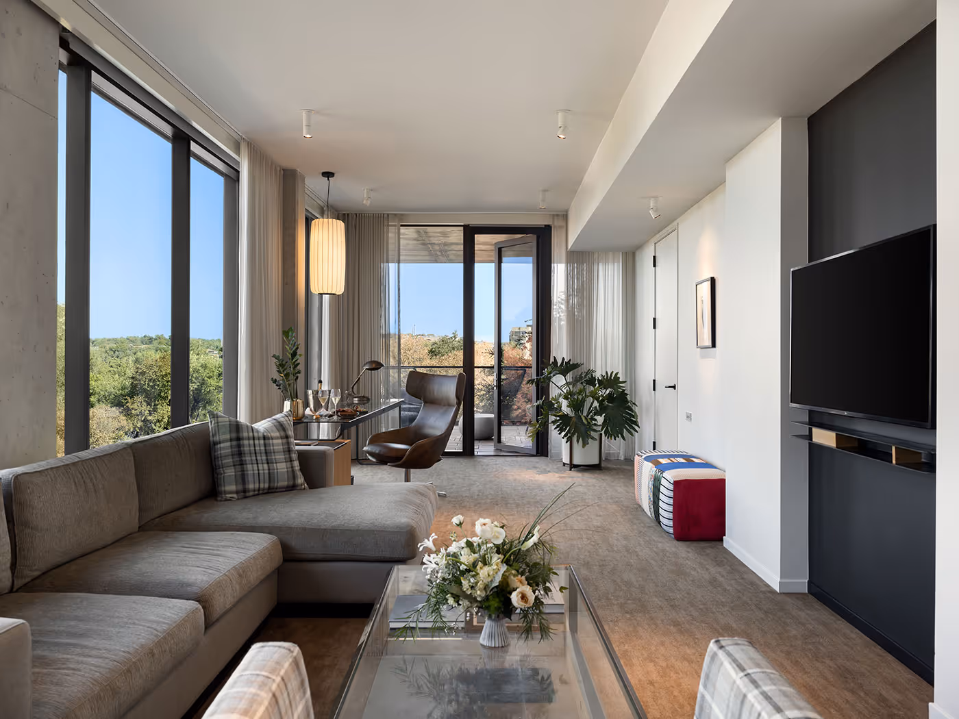Modern living room with large windows, an L-shaped sofa, coffee table, armchair, and wall-mounted TV.