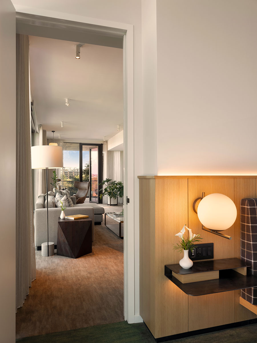 Modern hotel suite with a sitting area, neutral décor, floor-to-ceiling windows, and a lit wall sconce by the doorway.