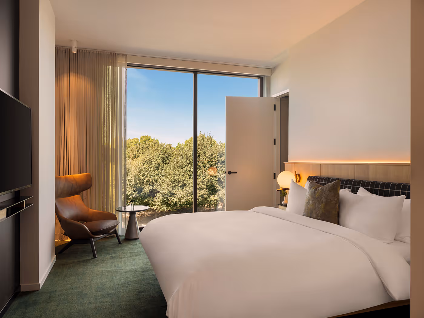 Modern hotel room with a large bed, armchair, TV, and floor-to-ceiling window overlooking trees outside.