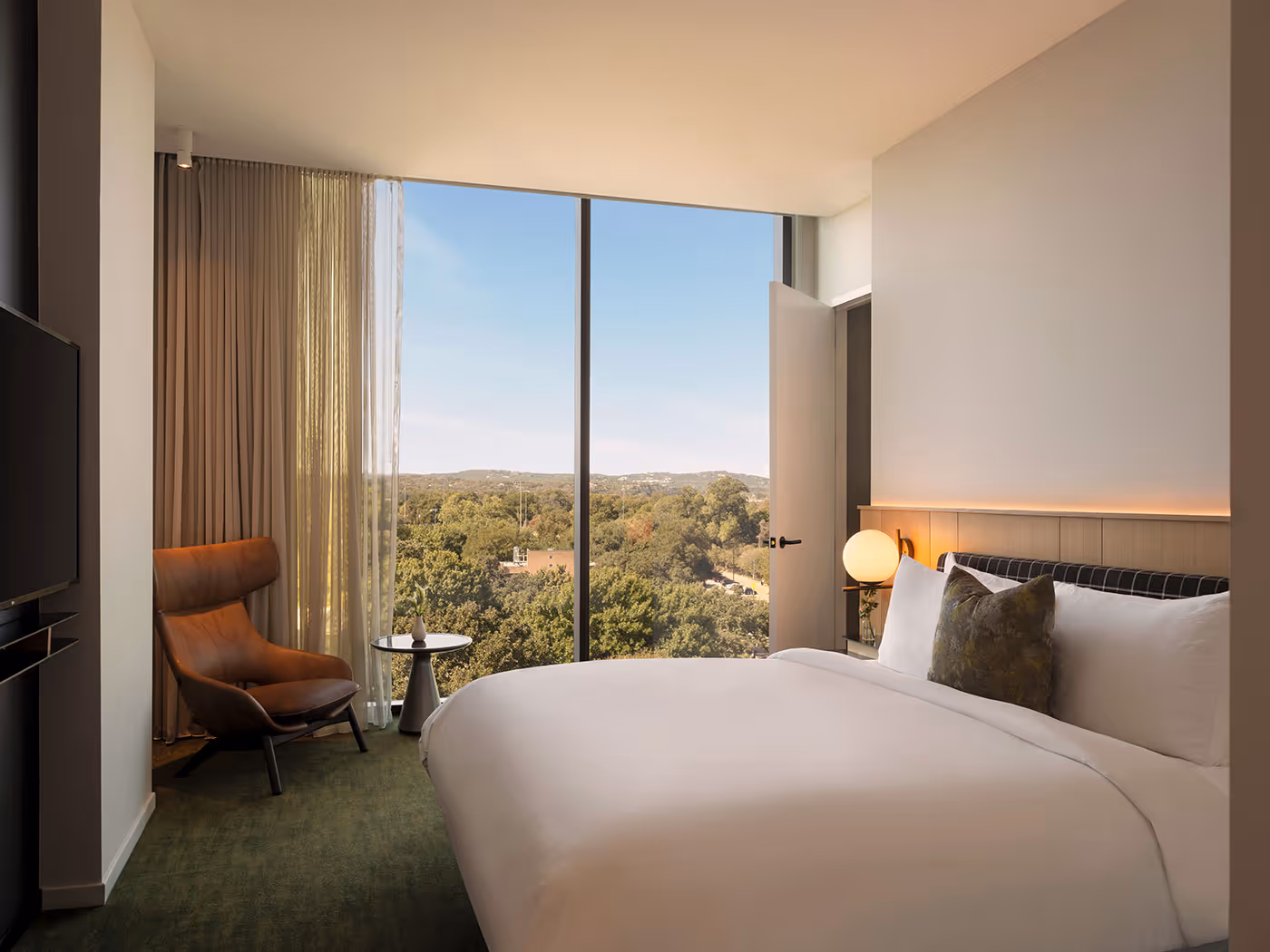 Modern hotel room with a large bed, armchair, floor-to-ceiling window, and view of trees and blue sky outside.