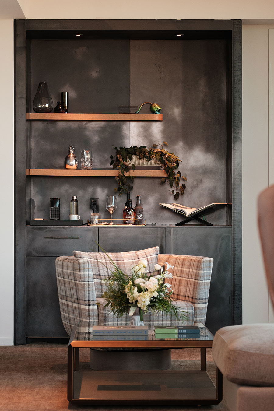 Modern lounge nook with plaid armchair, glass coffee table, and built-in shelves holding decor, books, and plants.