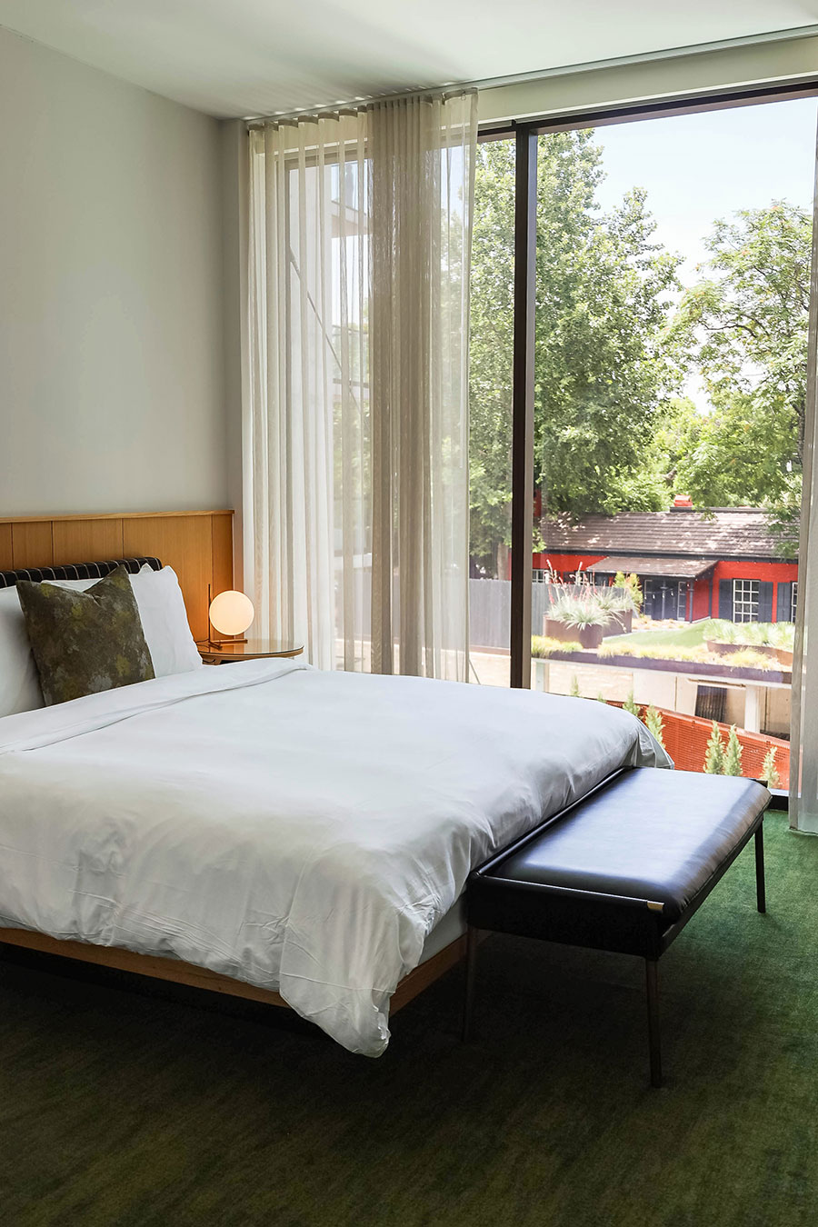 Modern bedroom with a large window, white bed linens, a small lamp, and a view of trees and a red building outside.