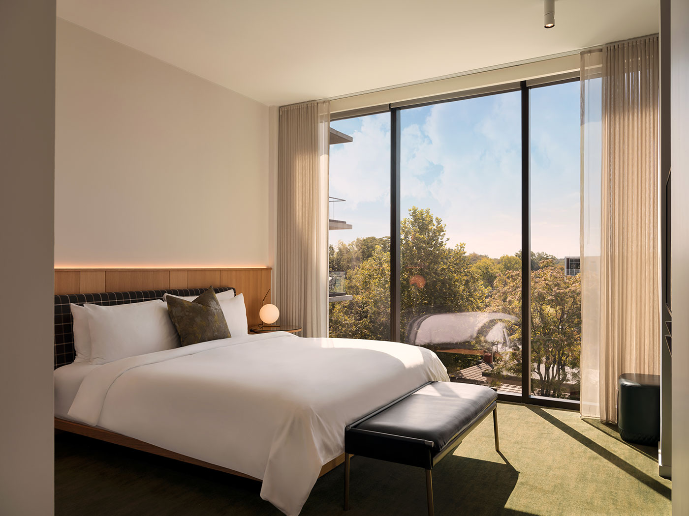 Modern hotel room with a large bed, bench, and floor-to-ceiling window overlooking trees and buildings under a blue sky.