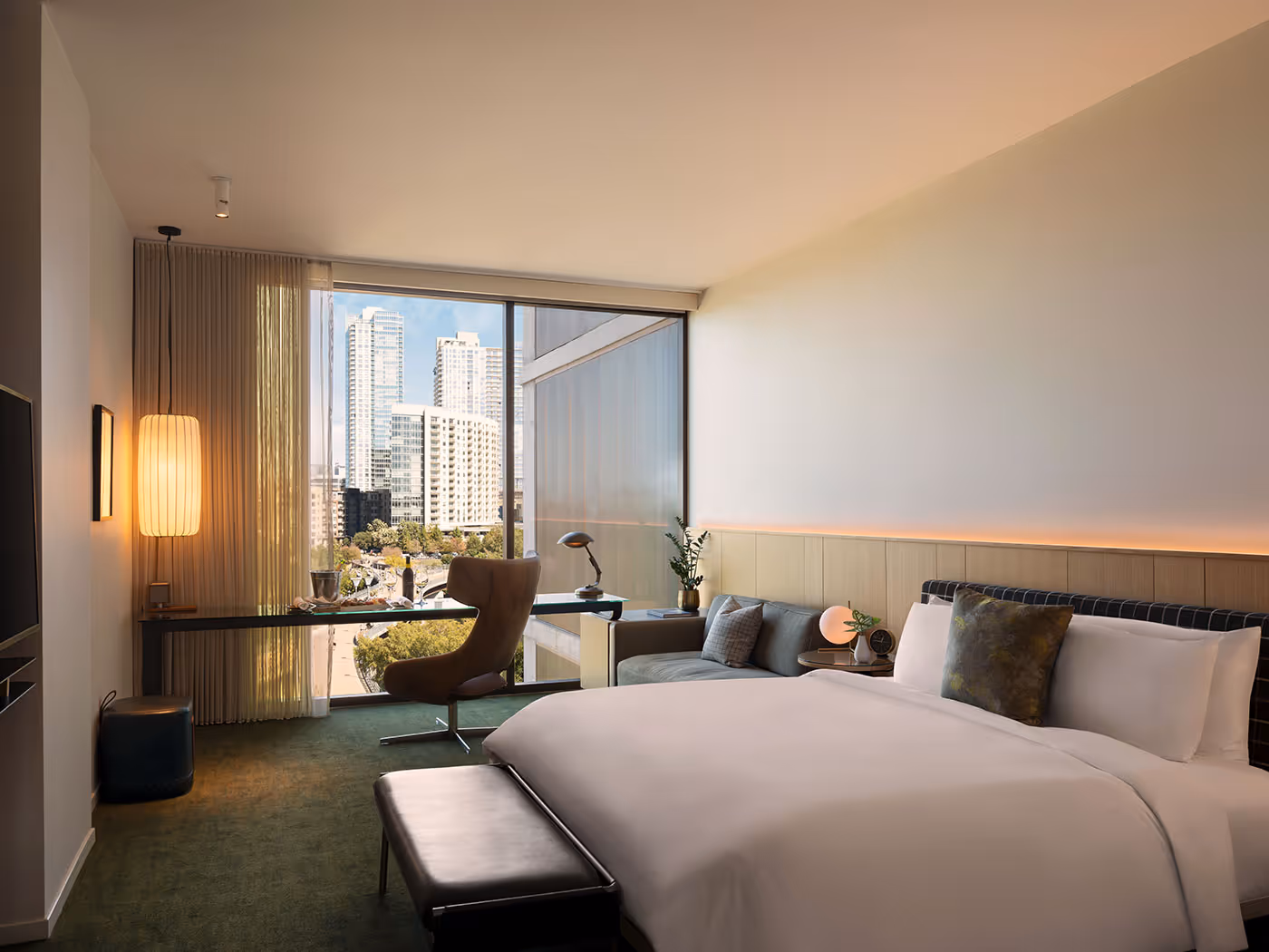 Modern hotel room with a large bed, desk, armchair, city view through floor-to-ceiling window, and neutral-toned decor.