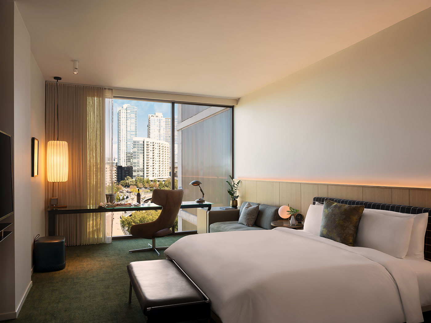 Modern hotel room with a large bed, desk, armchair, city view through floor-to-ceiling window, and neutral-toned decor.