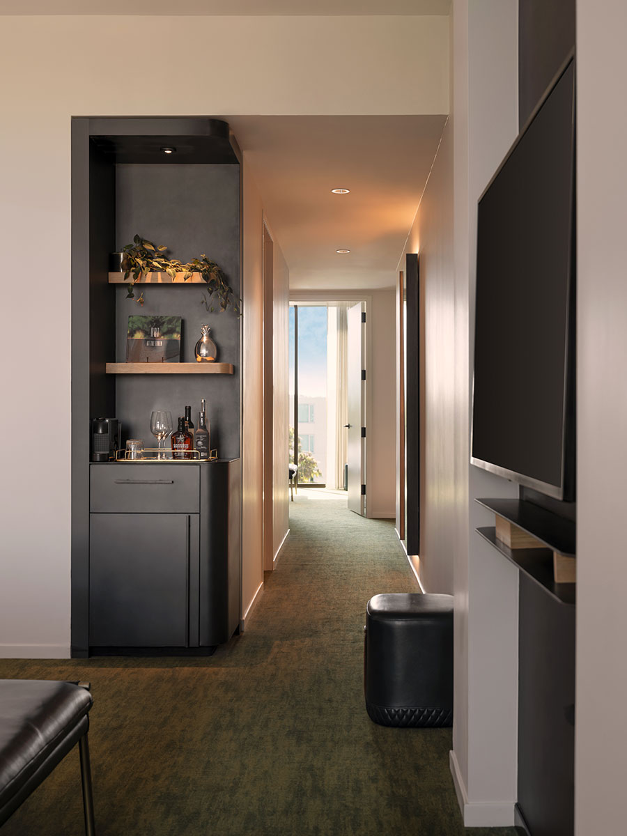 A modern hallway with a built-in bar, green carpet, and a view of sunlight through an open door at the end.