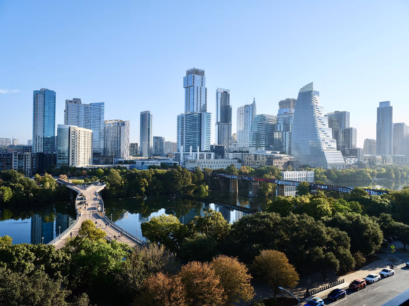 A city skyline with modern high-rise buildings, a river, and a pedestrian bridge surrounded by trees under a clear sky.