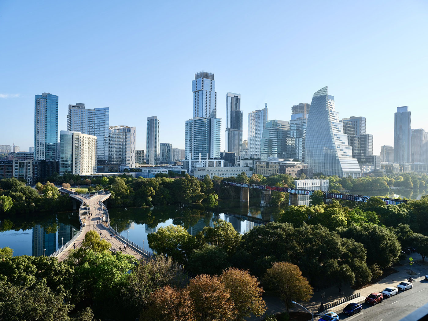 A city skyline with modern high-rise buildings, a river, and a pedestrian bridge surrounded by trees under a clear sky.