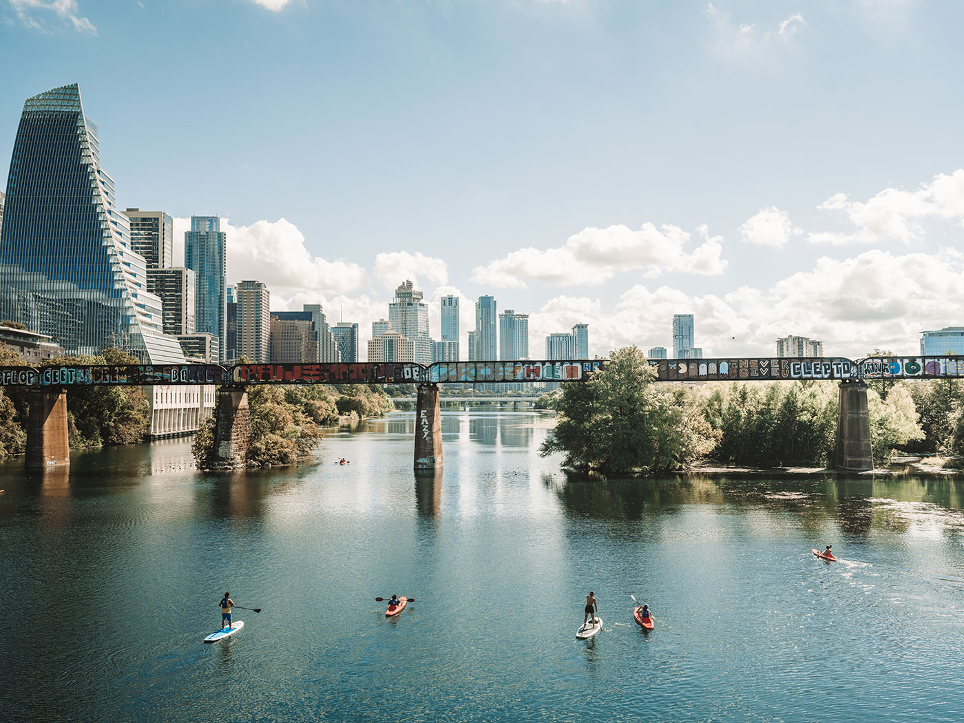 Paddleboarders and kayakers on a river with a graffiti-covered bridge and city skyline in the background.