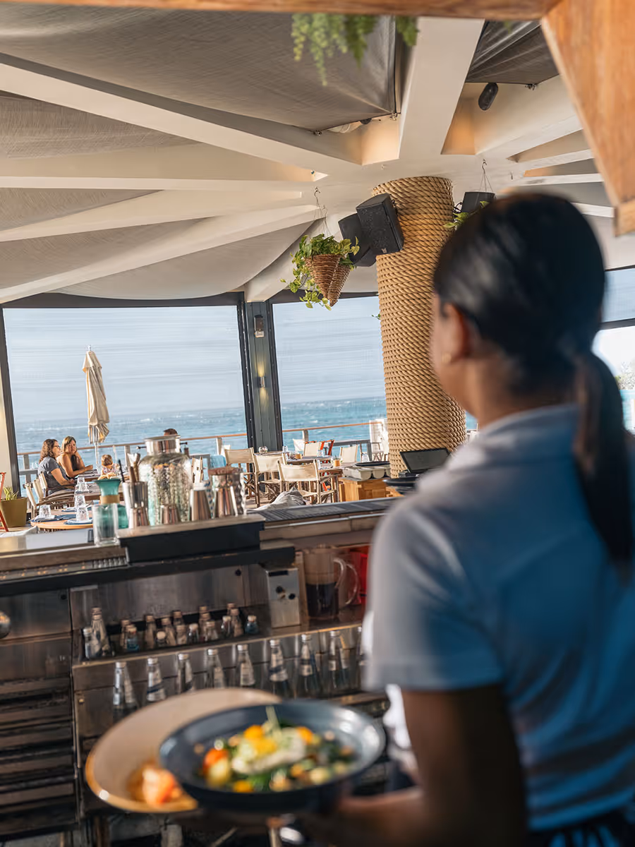A server holding a plate of food stands in an open-air restaurant with a view of the ocean and seated diners.
