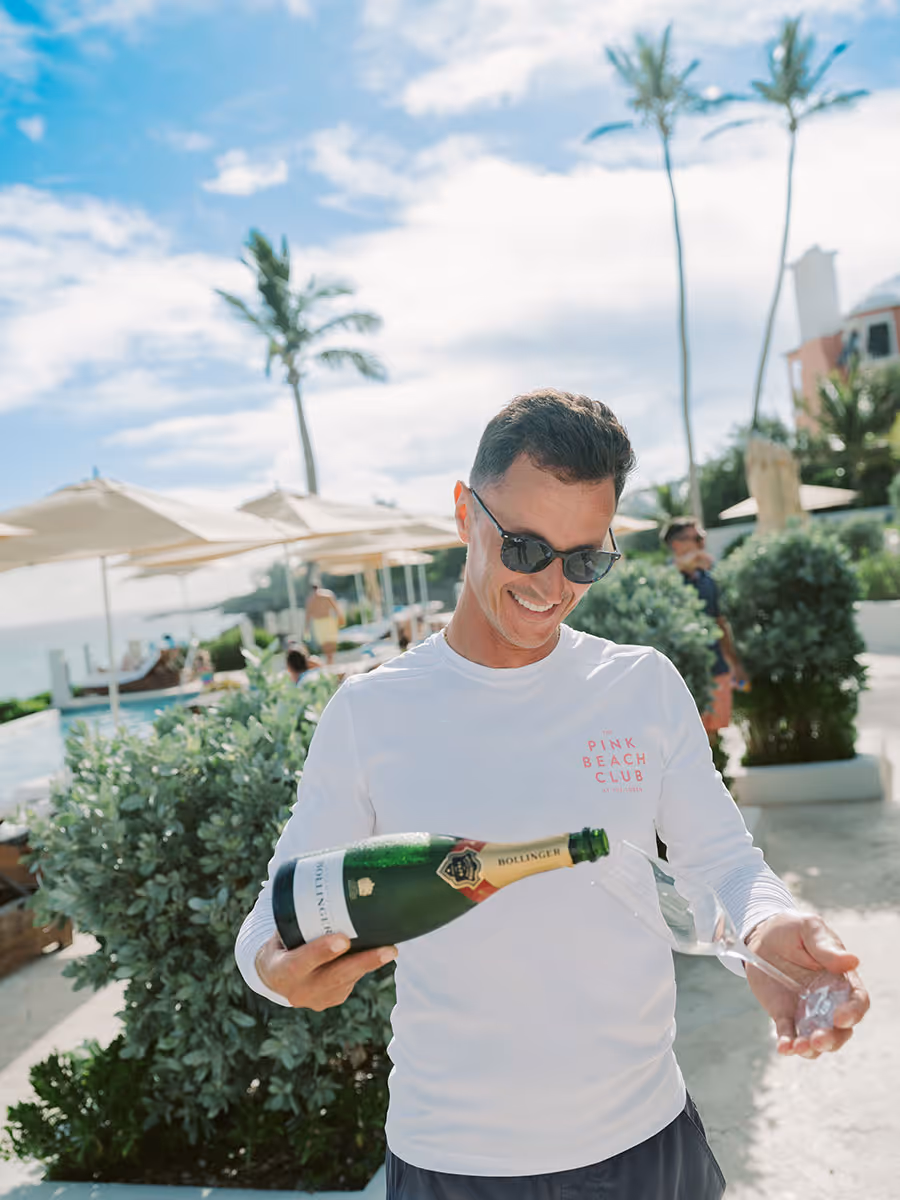 Man in sunglasses and white shirt pours champagne into a glass outdoors at a sunny, tropical resort.