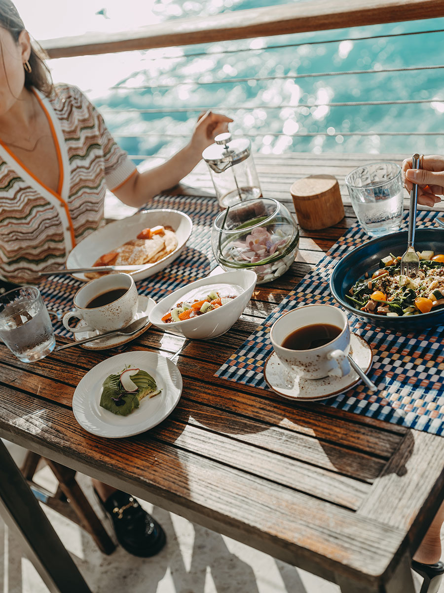 Two people dining outdoors by the water with plates of food, coffee cups, and glasses of water on a wooden table.