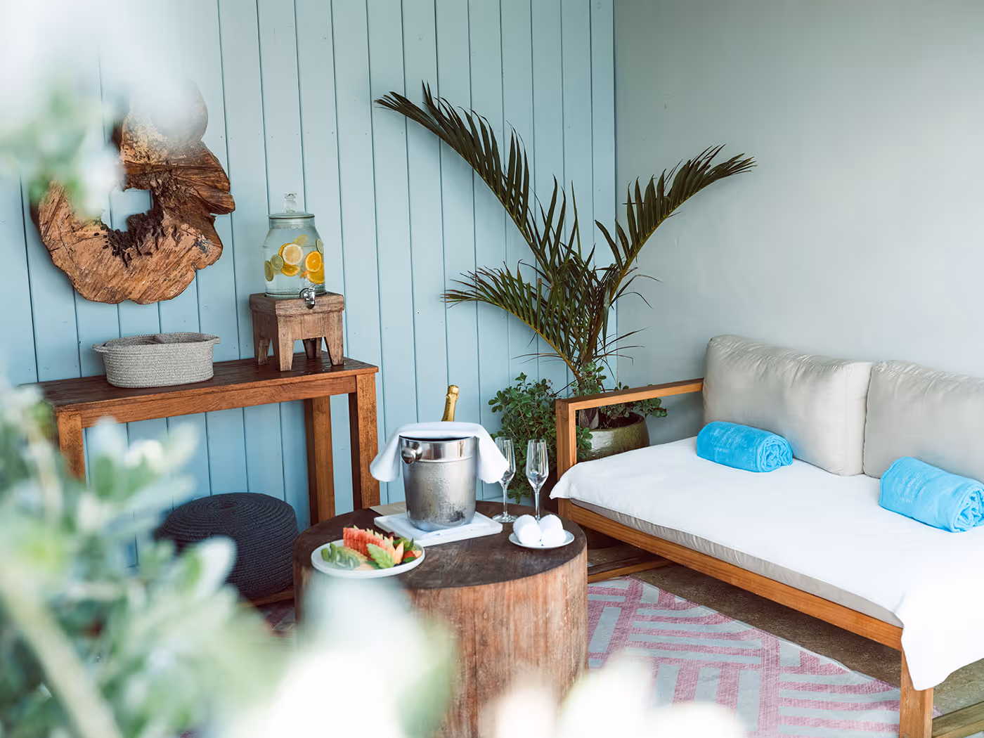 Outdoor seating area with a cushioned bench, wooden tables, a drink dispenser, a plant, and rolled blue towels.
