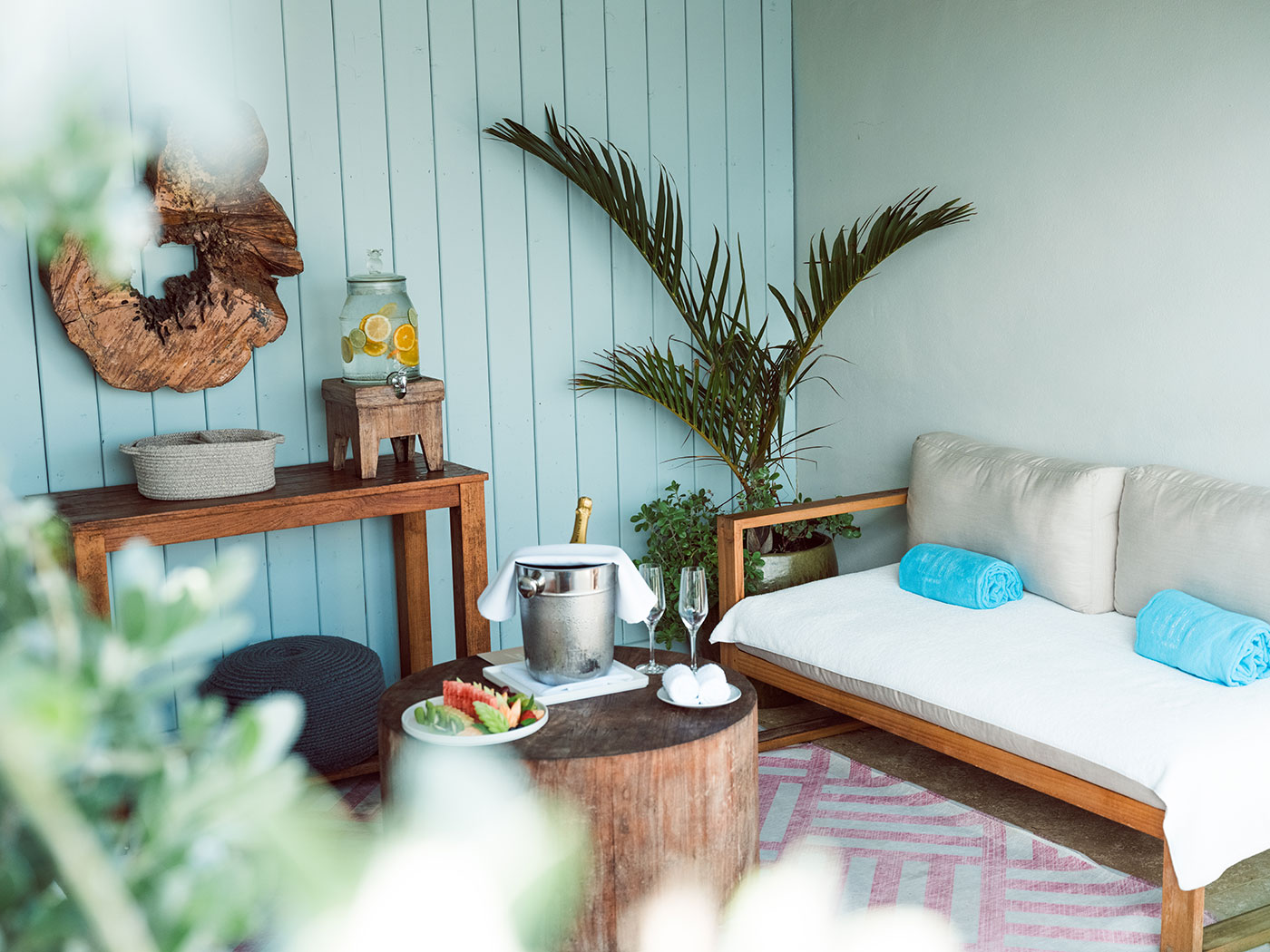 Outdoor seating area with a cushioned bench, wooden tables, a drink dispenser, a plant, and rolled blue towels.