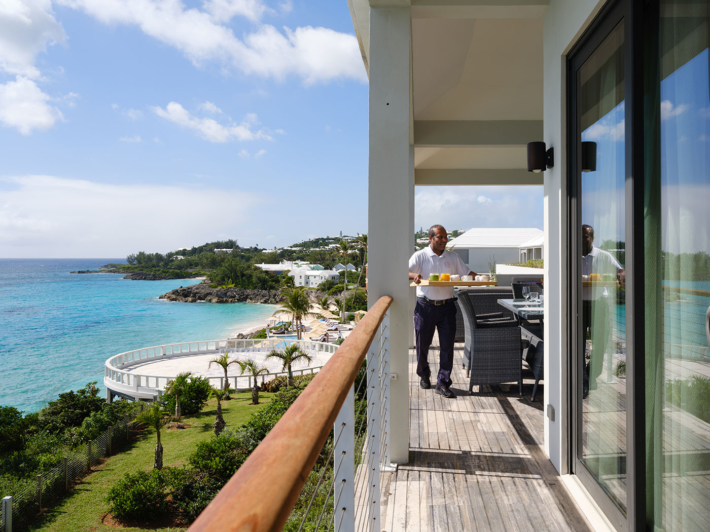 A man serves food on a balcony overlooking the ocean, tropical landscape, and white buildings on a sunny day.