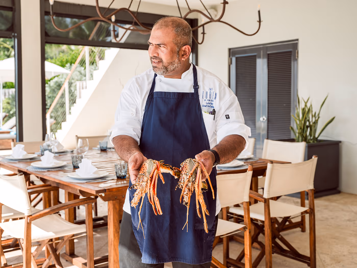 Chef in a blue apron stands indoors holding two large lobsters, with a set dining table in the background.