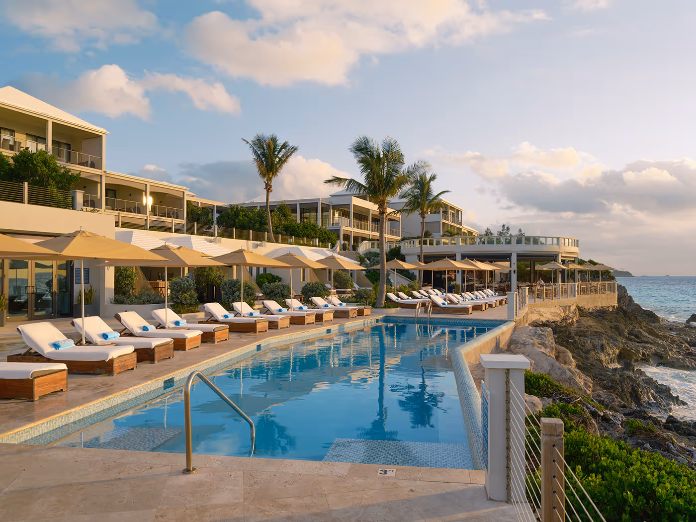 A resort pool with lounge chairs and umbrellas overlooks the ocean, surrounded by modern buildings and palm trees at sunset.