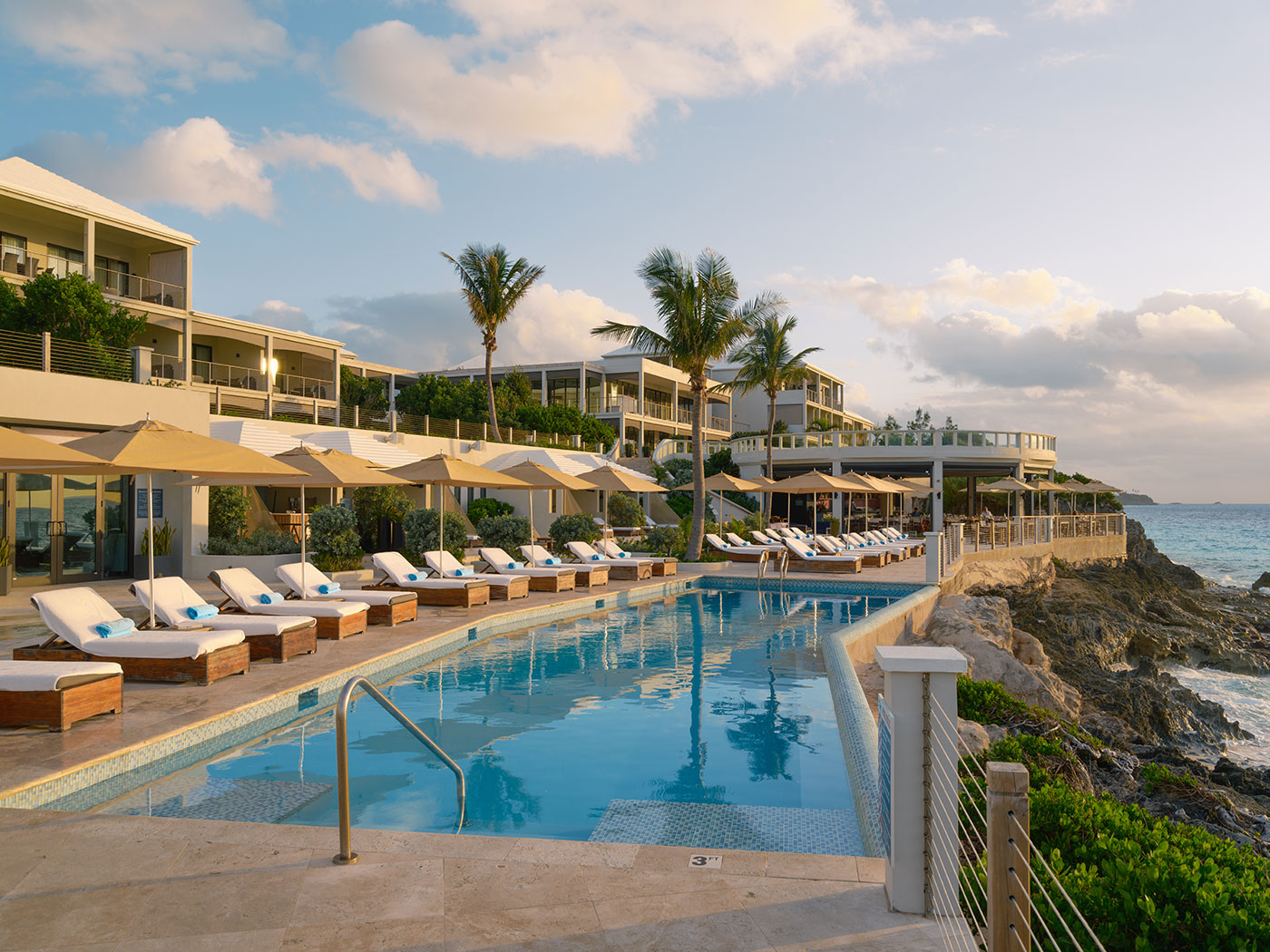 A resort pool with lounge chairs and umbrellas overlooks the ocean, surrounded by modern buildings and palm trees at sunset.