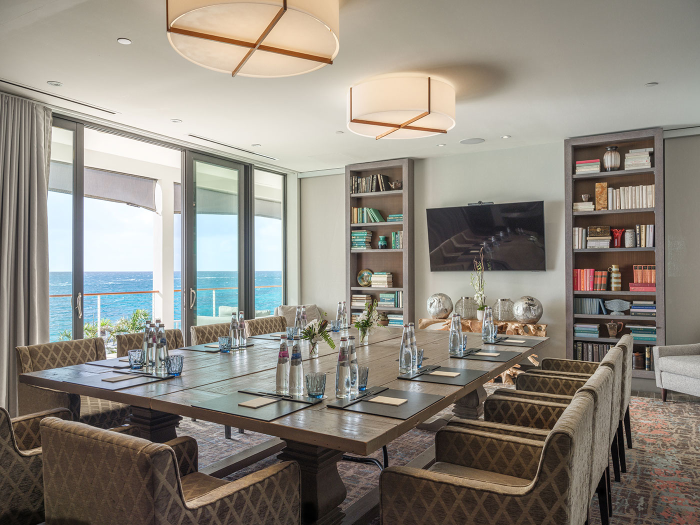A modern conference room with a large table, chairs, bottled water, notepads, TV, bookshelves, and an ocean view.