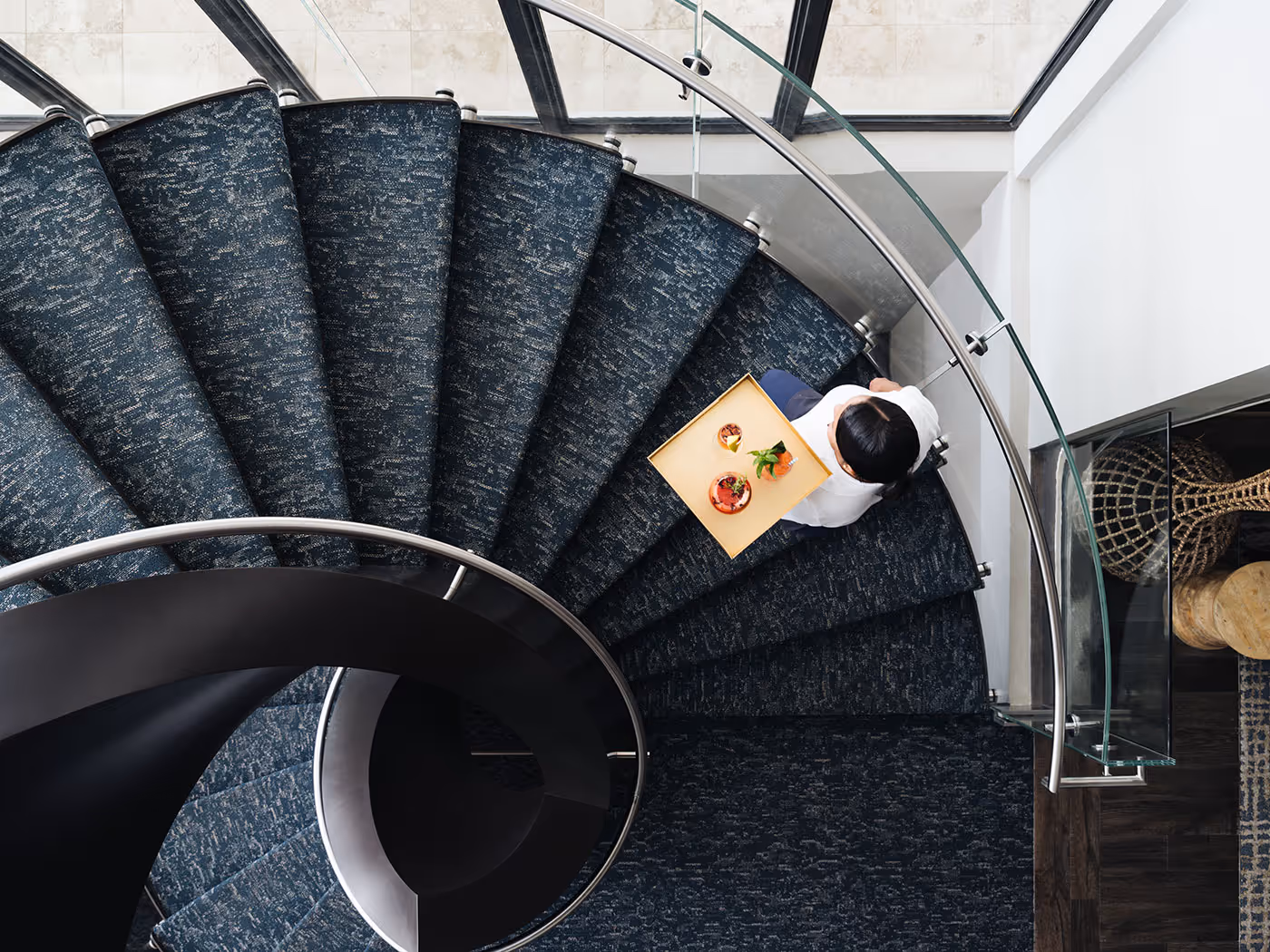 Person carrying a tray of food walking down a curved, carpeted staircase with glass railing, viewed from above.
