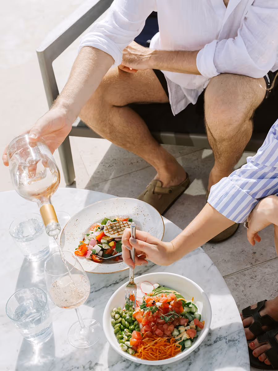 Two people sit at an outdoor table with salad bowls, pouring wine and reaching with a fork.