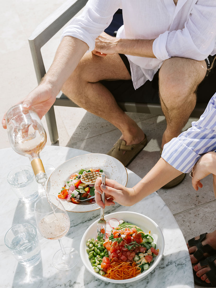 Two people sit at an outdoor table with salad bowls, pouring wine and reaching with a fork.