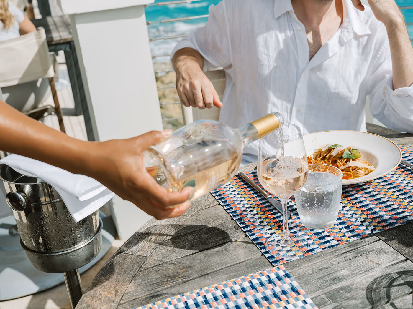 Server pouring white wine into a glass at an outdoor restaurant table with pasta and water on placemats.