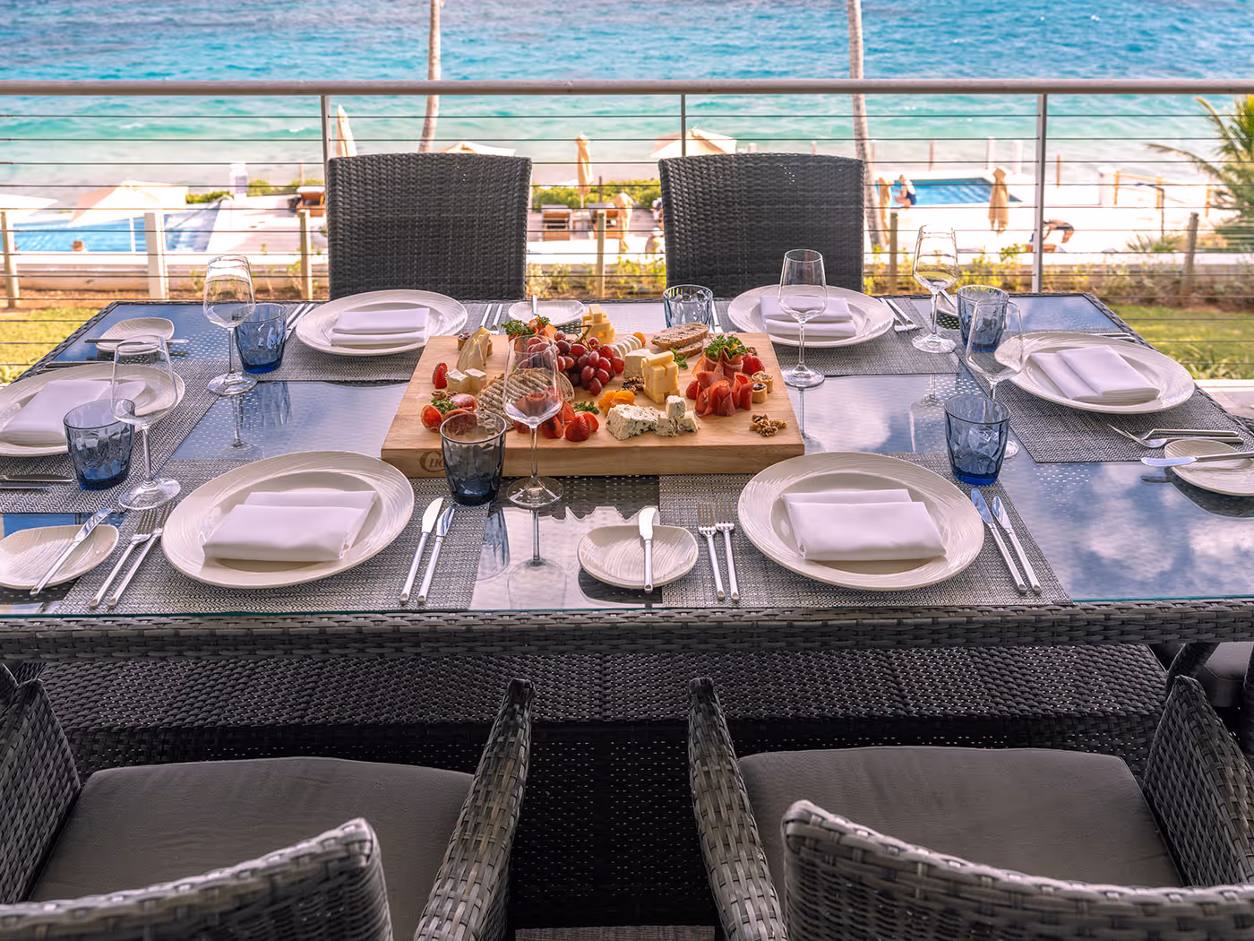 Outdoor dining table set for six with plates, glasses, and a cheese and fruit platter, overlooking the ocean.
