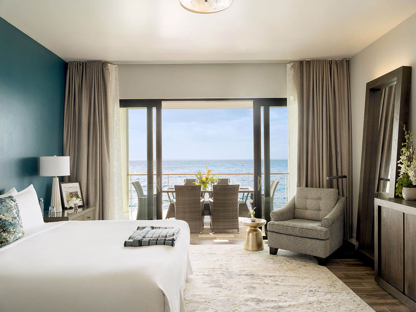 A hotel room with a bed, armchair, table, and a balcony overlooking the ocean under a clear sky.